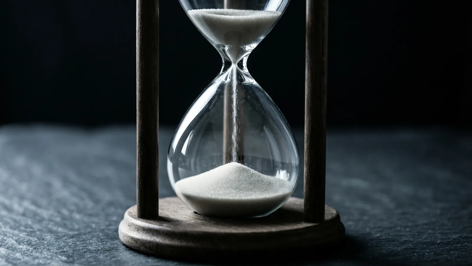 A glass hourglass in a dark wooden frame stands on slate against a black background, a thin thread of pale sand falling through the waist onto the pile in the lower bulb.