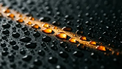 Hileras de gotas de agua redondas perlan una pizarra oscura; una única fuente de luz cálida enciende un destello brillante dentro de unas pocas, mientras la superficie que las rodea permanece en sombra profunda.