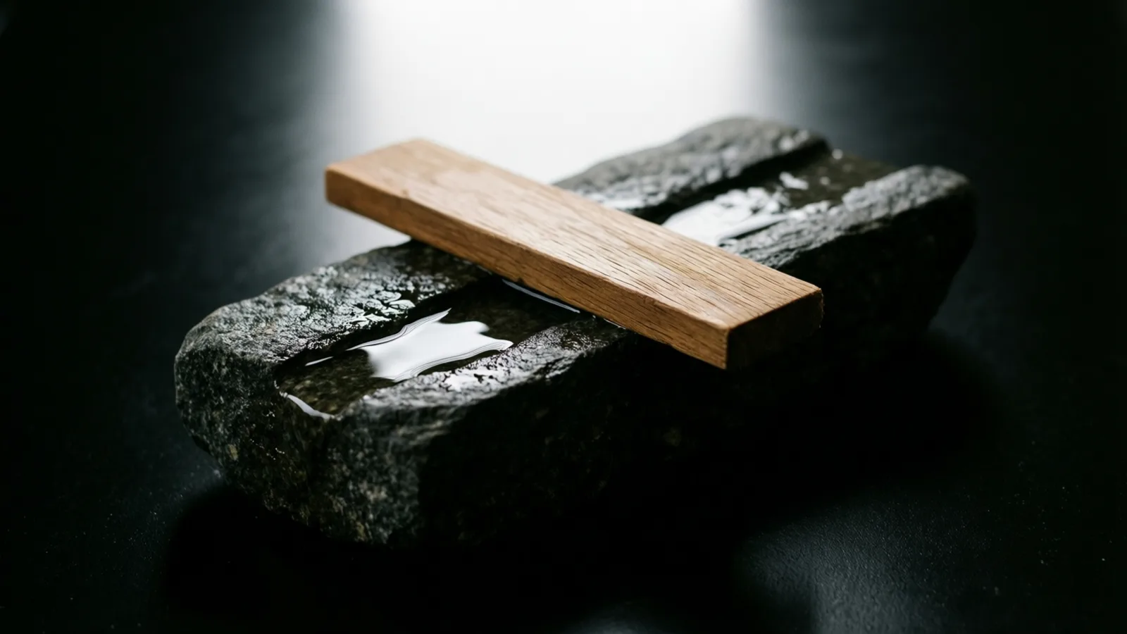 Una tira plana de madera clara cruza una piedra oscura y áspera, con pequeños charcos de agua en las hendiduras de la roca. La superficie de abajo es negra y brillante, y una luz intensa llena el fondo desenfocado en la parte superior.