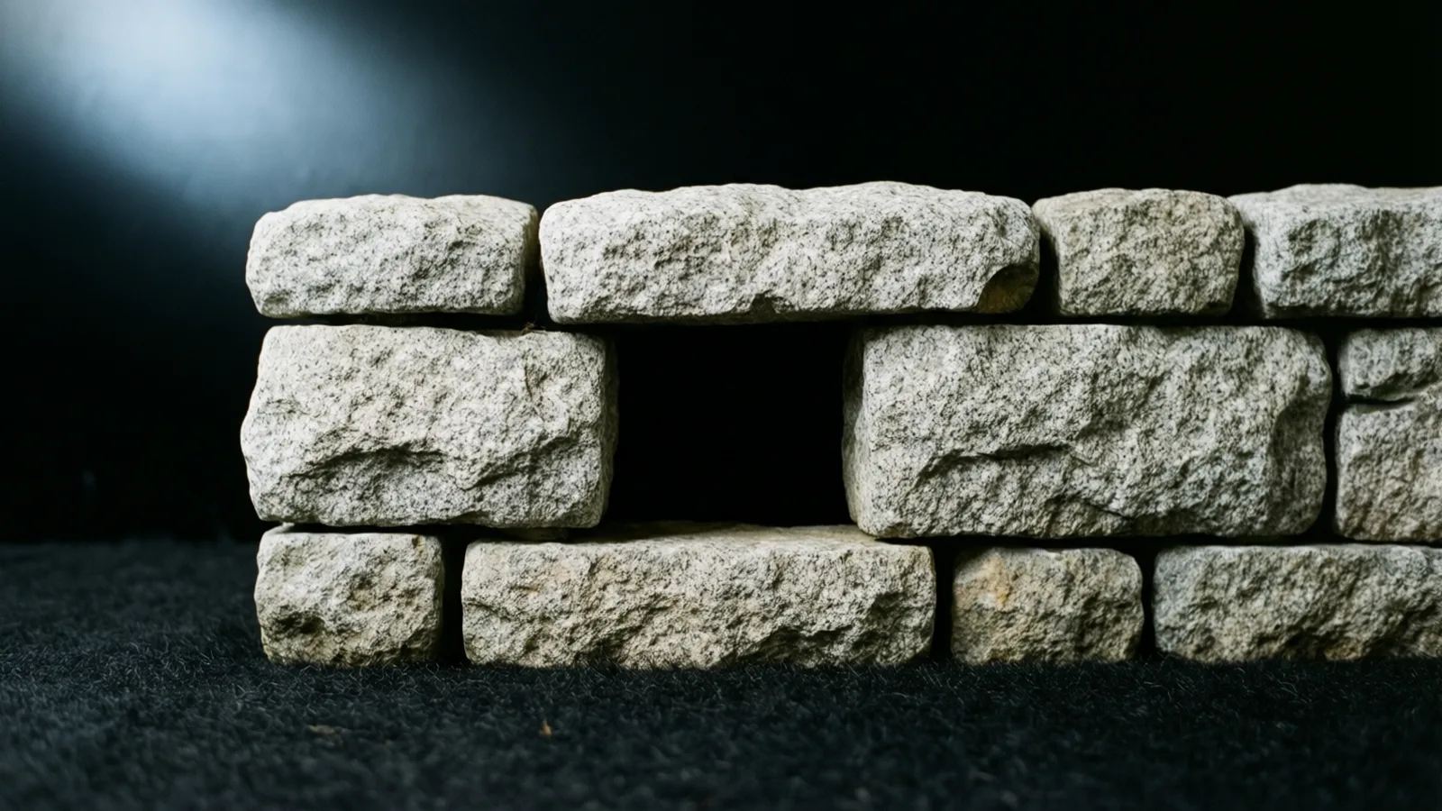 Un muro bajo de bloques de piedra rugosa, gris claro y moteada, con un hueco rectangular oscuro en el centro, sobre una tela negra de aspecto de alfombra. La luz suave cae sobre el frente de los bloques, con un resplandor pálido en el fondo oscuro, arriba a la izquierda.