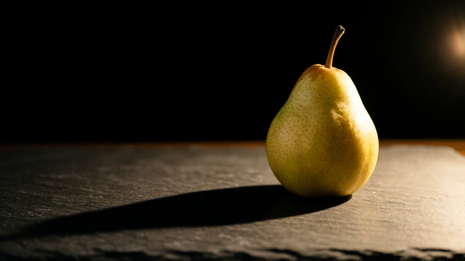 Una sola pera verde amarillenta con el tallo curvo está sobre una superficie de pizarra oscura. La luz llega desde la derecha, ilumina la fruta y proyecta una sombra larga hacia la izquierda, con fondo negro.