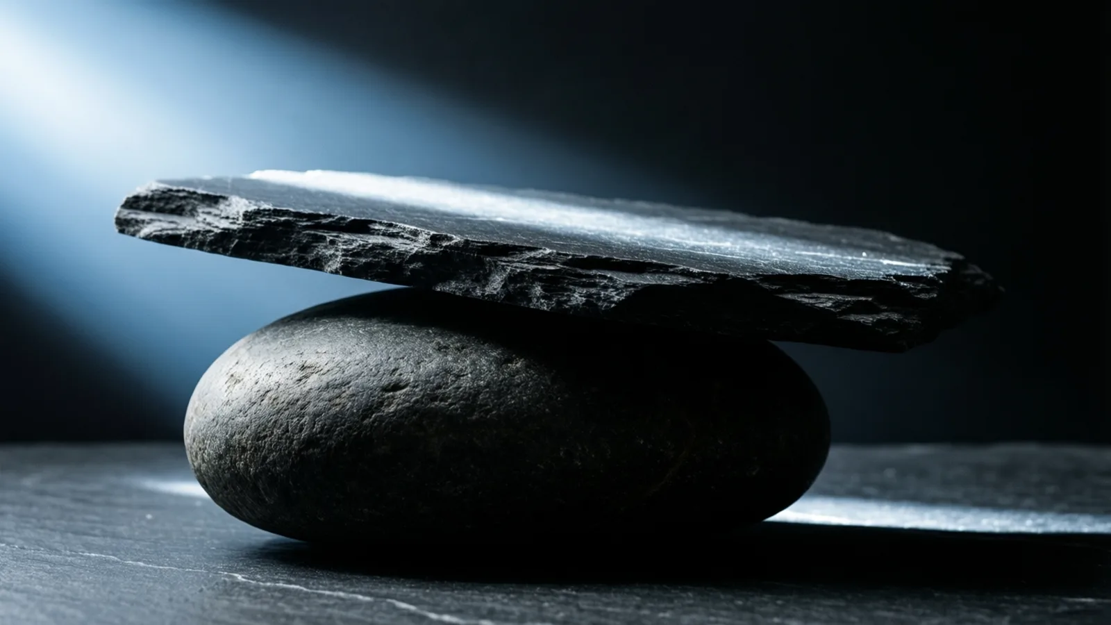 A thick slate slab with a rough fractured edge rests tilted across a smooth rounded dark stone on a scratched slate surface, a single cold blue beam raking in from the upper left.