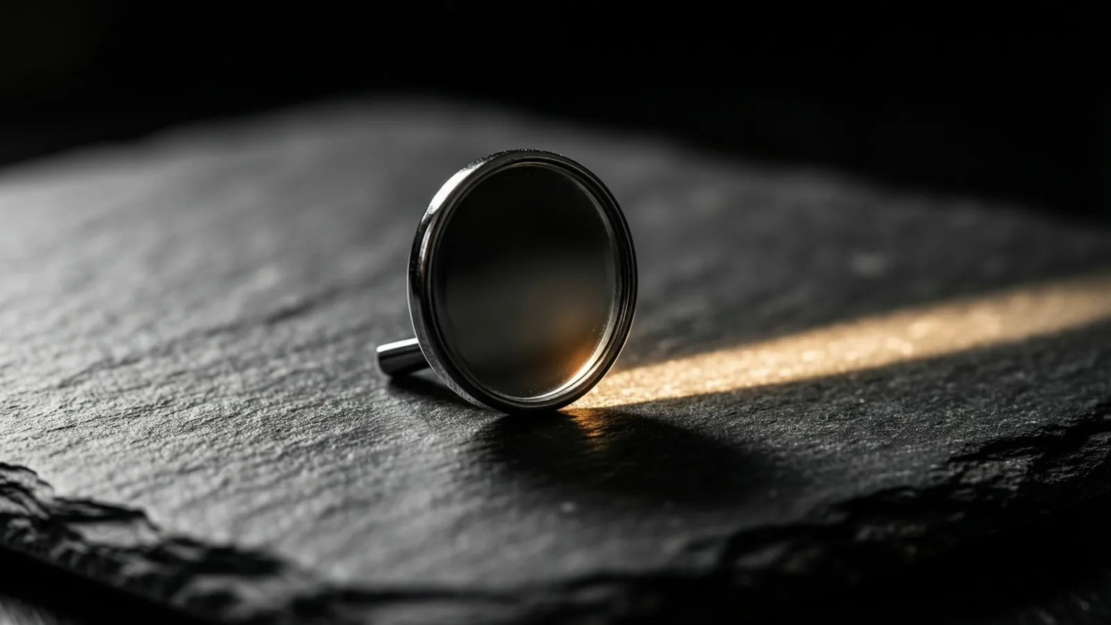 A small round mirror in a polished steel rim stands tilted on dark slate, a faint warm glow low on its face, while a band of warm light crosses the stone and its shadow falls across that band.