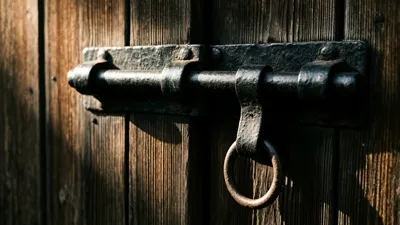 A heavy dark iron sliding bolt is drawn across weathered vertical wooden boards, a worn iron ring hanging from its handle, with warm light raking in from the left.