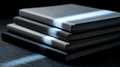 Four closed ledgers of different heights sit stacked unevenly on dark slate, cold light tracing the stair-step edges of their covers.