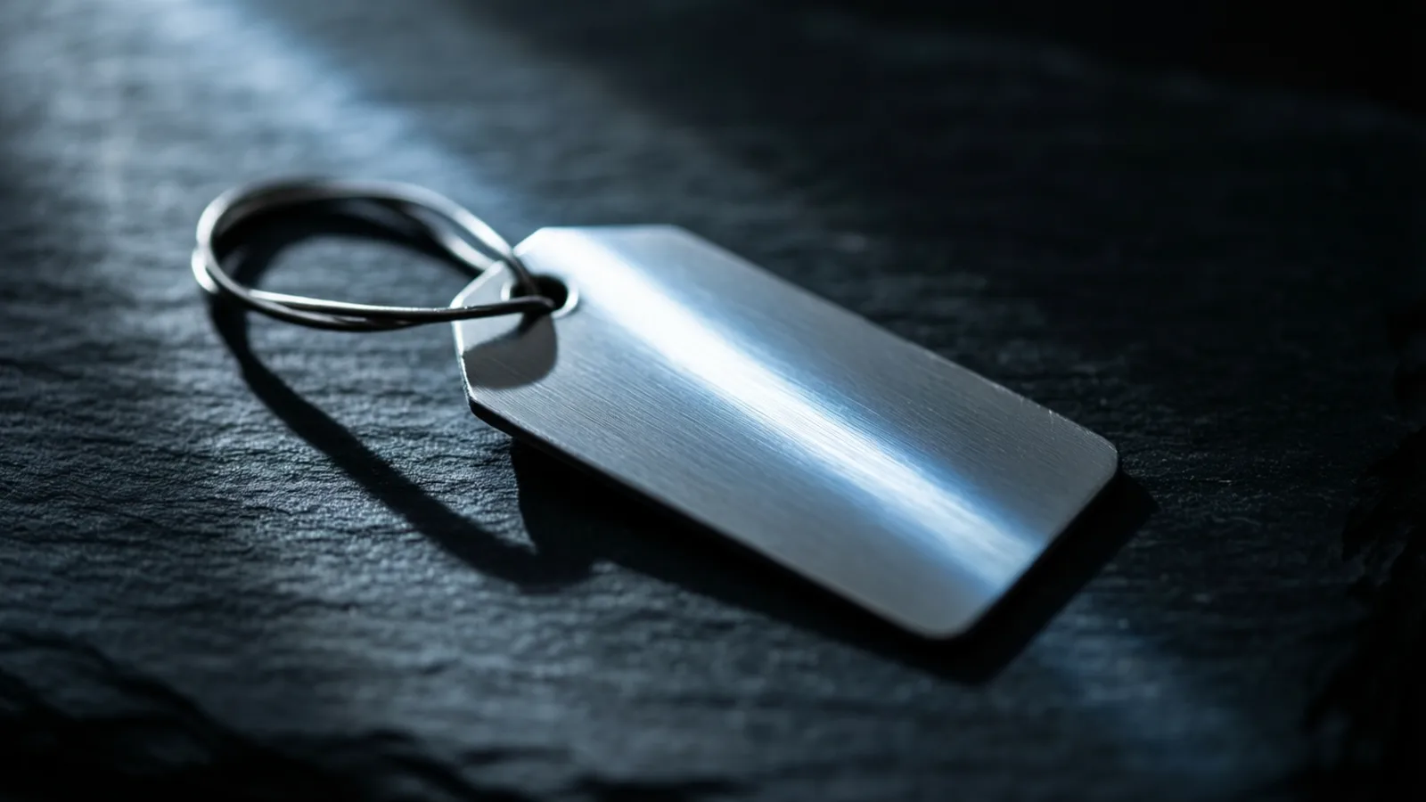 Una pequeña placa metálica sin inscripción, tumbada sobre pizarra oscura junto a un lazo enrollado de alambre fino, con un canto que prende una línea de luz fría.