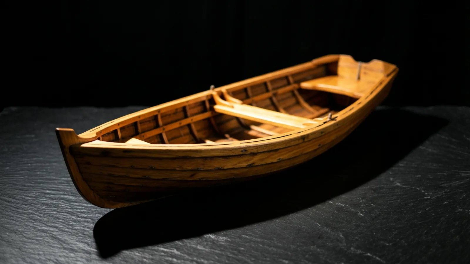 Un pequeño modelo de bote de remos de madera, con bancos abiertos y cuadernas en su interior, descansa sobre una superficie de pizarra gris oscuro. Una luz cálida cae desde arriba sobre el bote y proyecta una larga sombra hacia abajo a la derecha sobre un fondo negro.