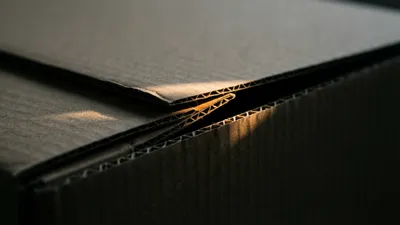 A cardboard carton's interlocking flap folded shut, its tabs lit by a single warm band of light against dark slate.