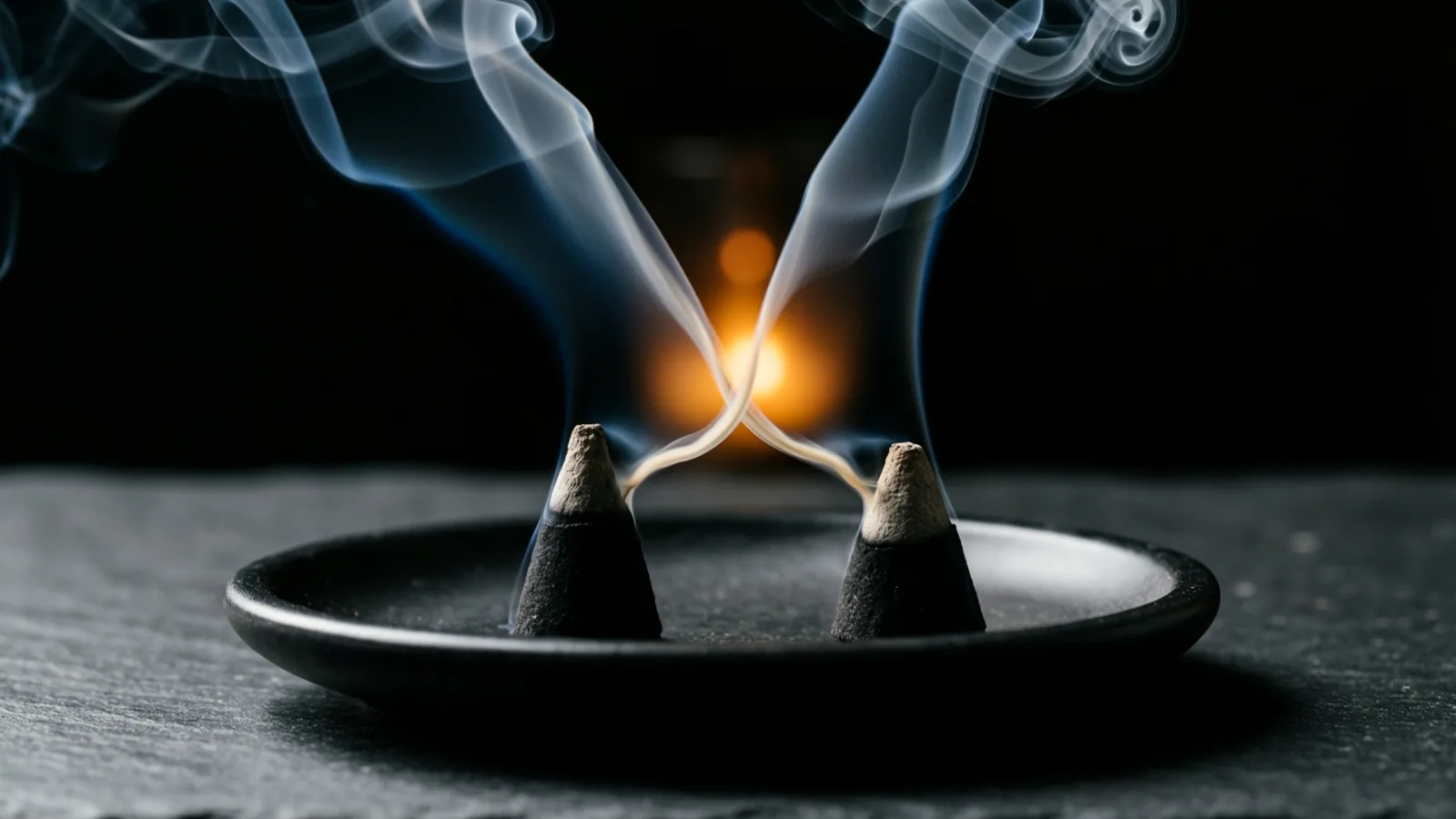 Dos conos de incienso oscuros con la punta clara descansan sobre un plato negro y redondo, sobre una superficie oscura; de cada uno sube humo que se enrosca y se cruza en el centro. La luz toca el humo y las puntas, y detrás hay un resplandor naranja desenfocado sobre fondo negro.
