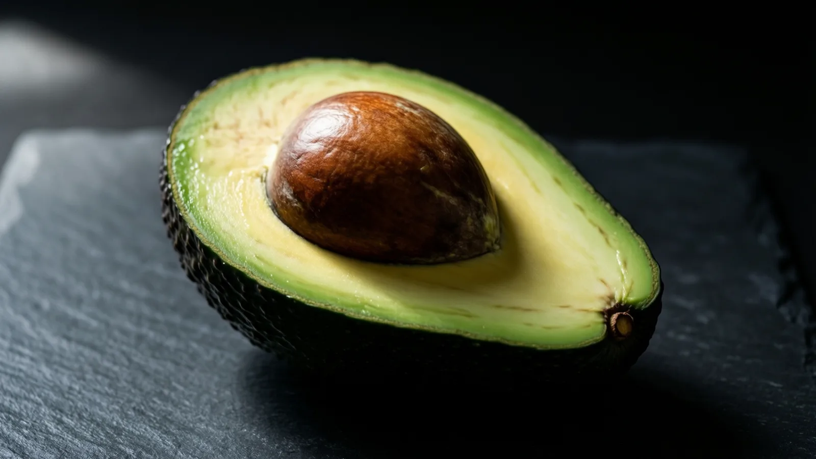 Medio aguacate con el hueso café liso a la vista, apoyado con el corte hacia arriba sobre una losa de pizarra oscura. La luz llega desde arriba a la izquierda sobre la pulpa verde pálido y el hueso brillante; el fondo es negro.