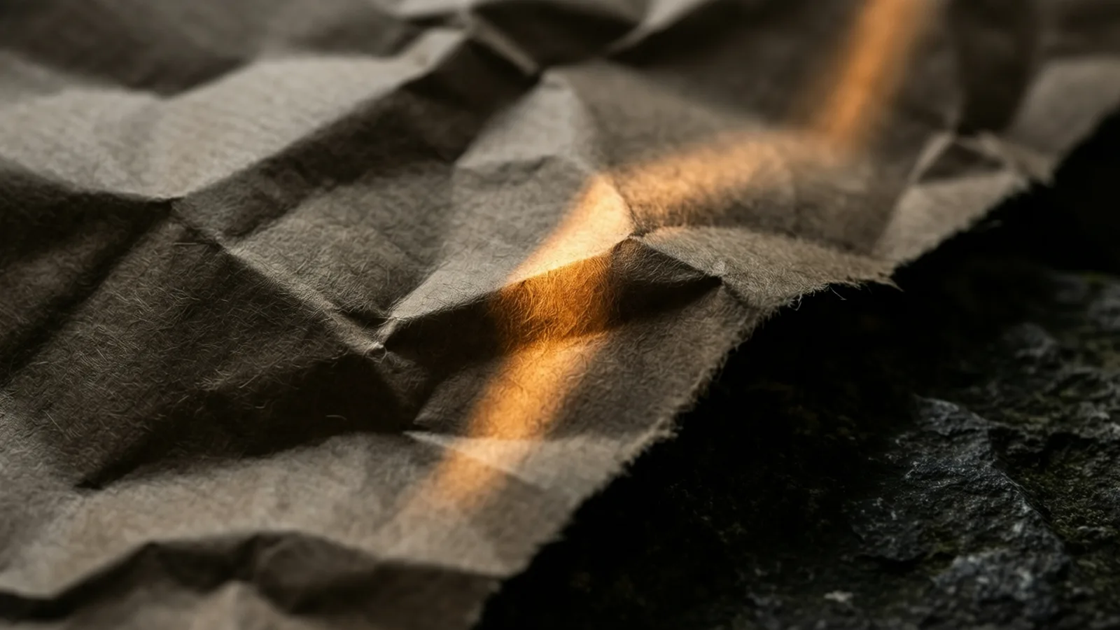 Crumpled kraft packing paper with deep folded creases, one warm band of light catching a raised fold against dark stone.