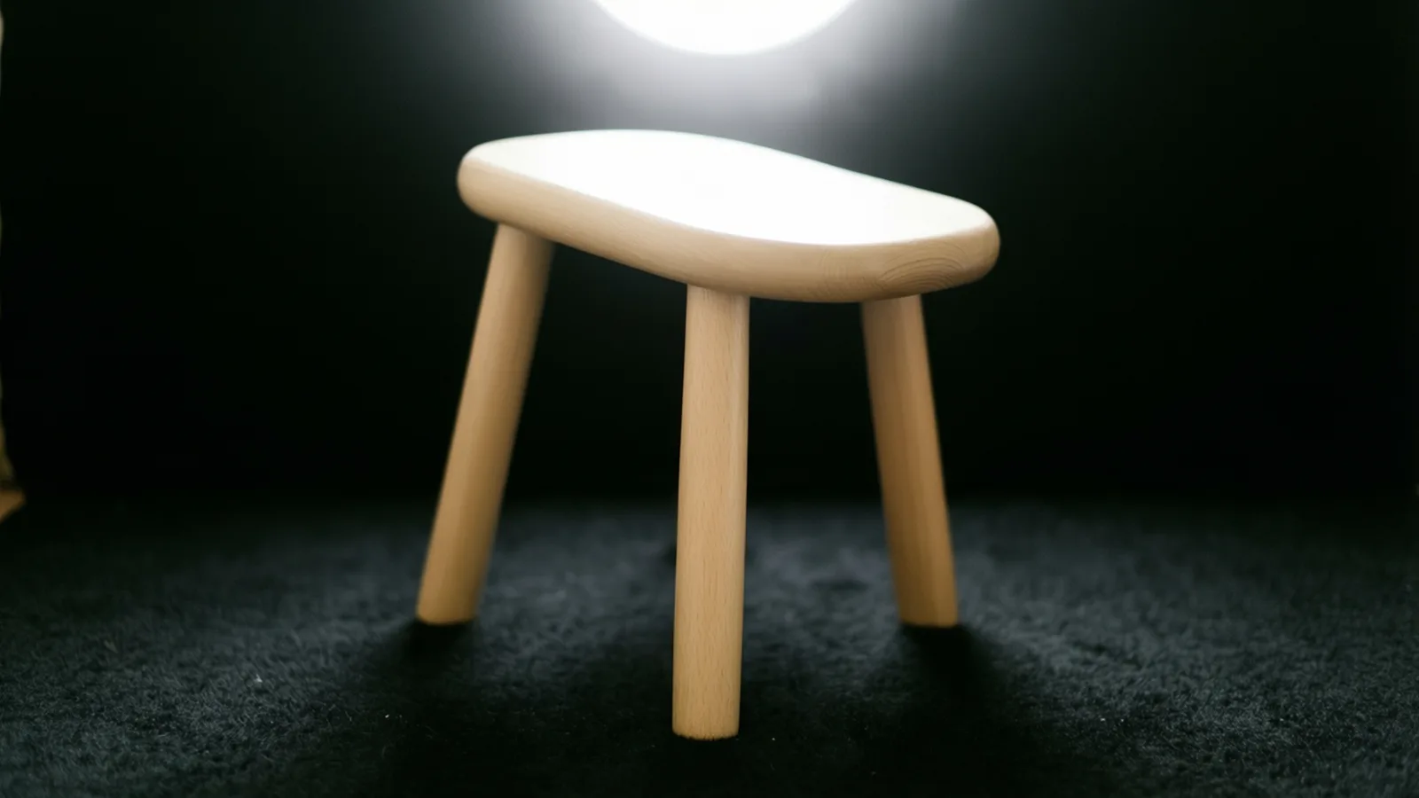Un taburete de madera clara, con asiento oblongo de bordes redondeados y patas redondas y delgadas, está sobre una alfombra oscura de pelo largo. Una luz redonda y brillante detrás y arriba borra el detalle de la parte superior del asiento, contra un fondo negro.