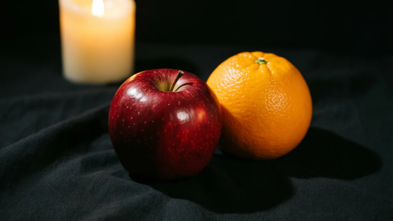 Una manzana roja brillante y una naranja descansan juntas sobre una tela negra arrugada. La luz llega desde la izquierda y resalta la piel de la manzana, con sombras suaves hacia la derecha. Al fondo, una vela crema desenfocada con una pequeña llama sobre un fondo oscuro.