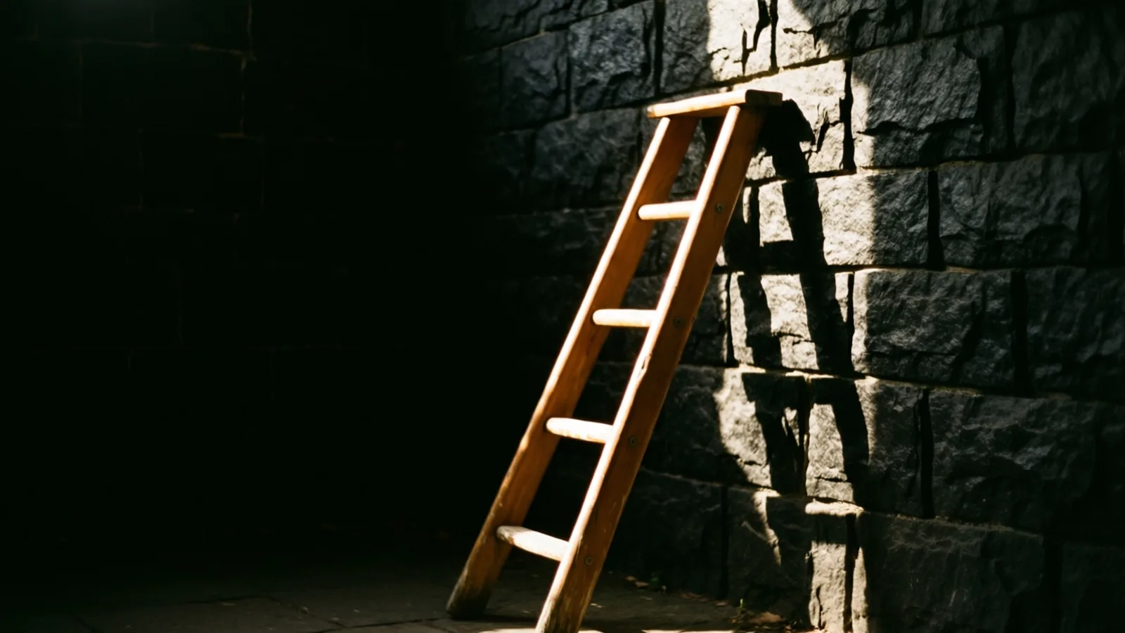 Una escalera de madera con unos pocos peldaños apoyada contra un muro de bloques de piedra oscura y rugosa. La luz del sol cae sobre la escalera y la parte derecha del muro, proyectando su sombra en las piedras, mientras el lado izquierdo queda casi negro.