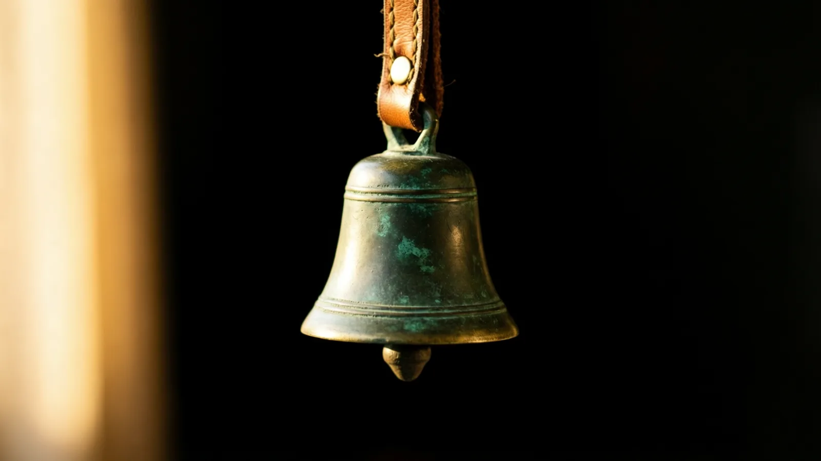 Una campana pequeña de metal envejecido, con pátina verde, cuelga de una correa de cuero café claro con un remache pálido. La luz resalta el hombro y el borde, y un resplandor cálido desenfocado ocupa el lado izquierdo sobre fondo negro.