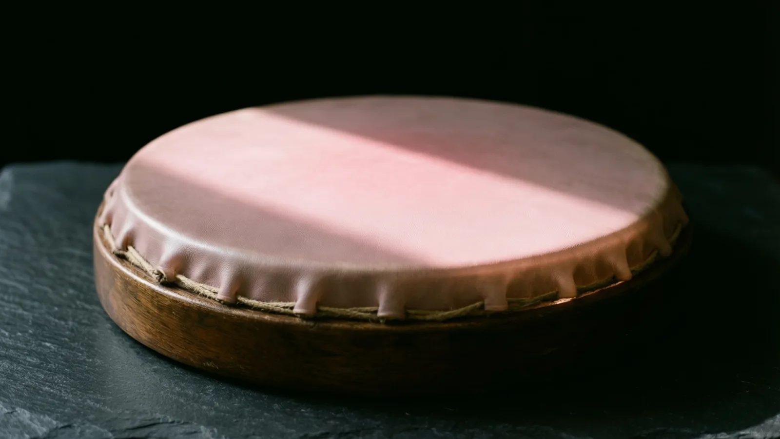 A round, shallow drum-like object with a smooth pink leather top stitched at the edge with cord, on a dark wooden rim, resting on grey slate. A band of light crosses the top against a black background.