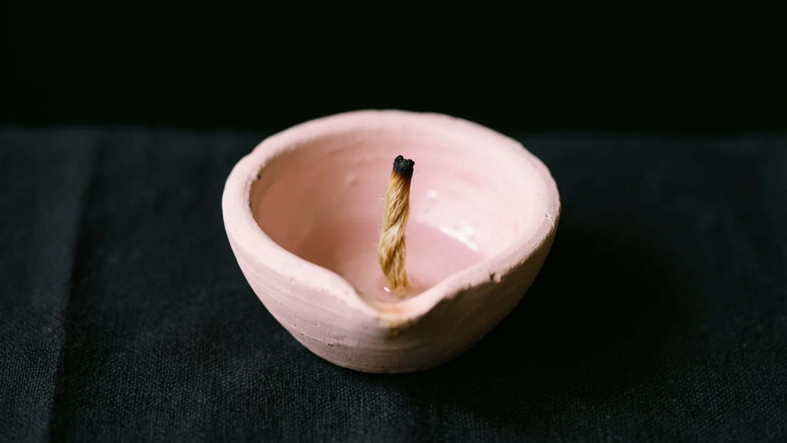 Un cuenco pequeño de cerámica rosa y rugosa descansa sobre una tela oscura, con un cordón corto de fibra natural retorcido que se alza en su interior y tiene la punta ennegrecida. La luz cae sobre el cuenco y el cordón, y deja la tela y el fondo negro en la oscuridad.