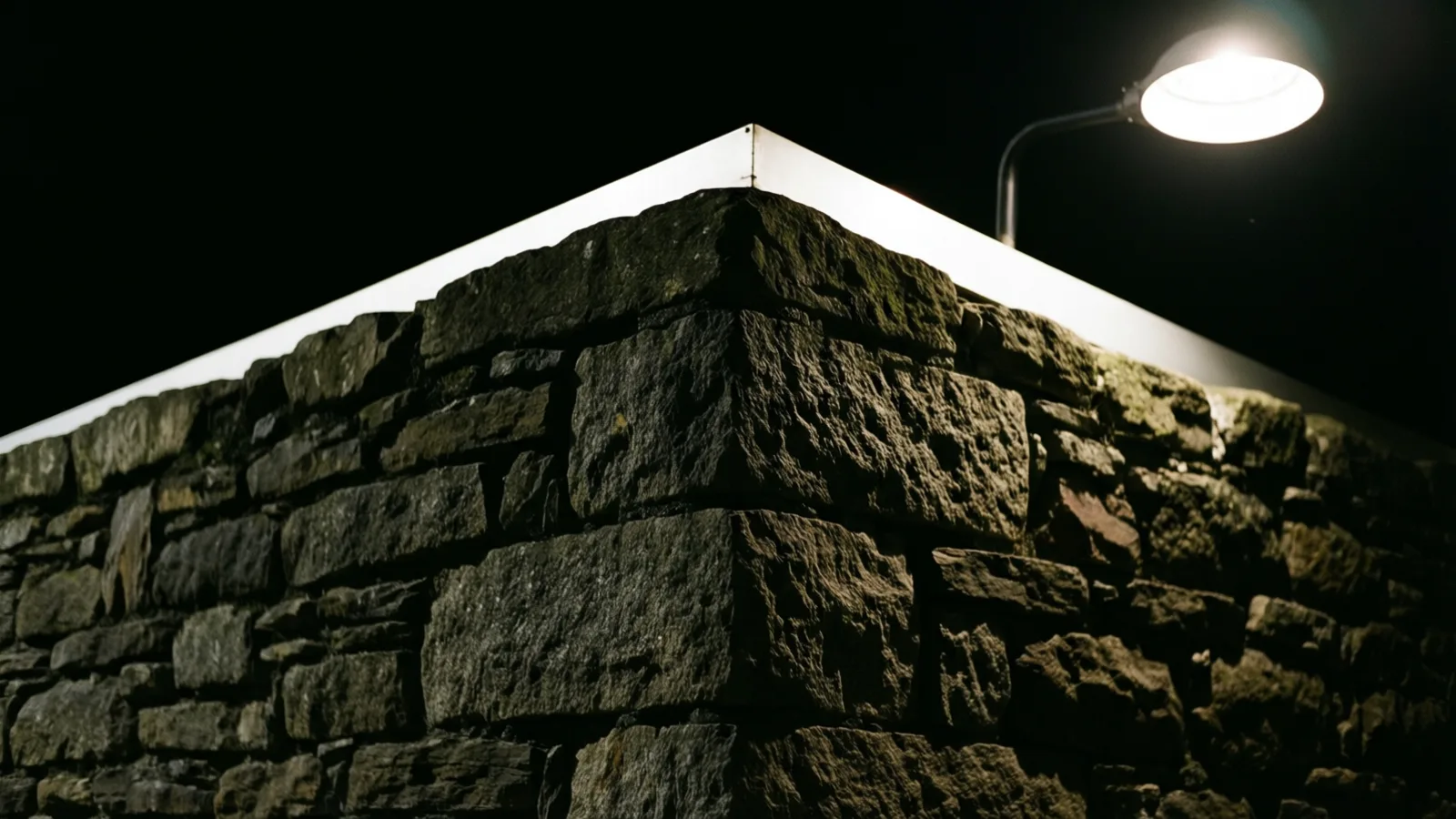 La esquina de un muro rugoso de piedra seca vista desde abajo, rematada por una cubierta blanca en ángulo. Una lámpara redonda sobre un brazo curvo, arriba a la derecha, brilla intensamente contra un cielo nocturno negro.