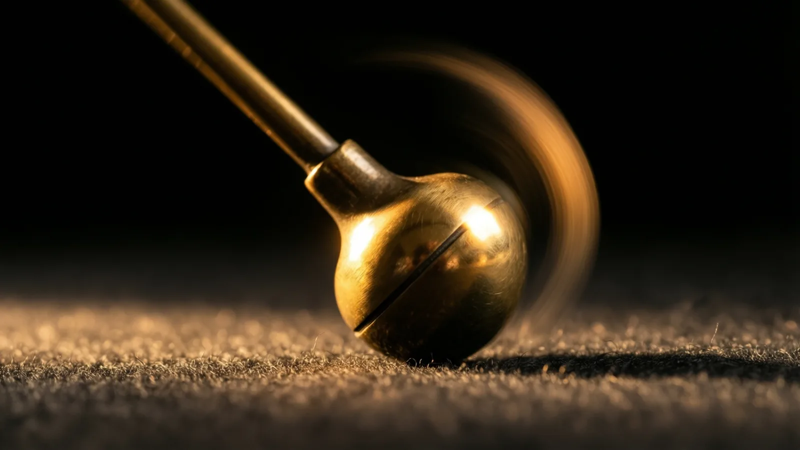 Una lenteja de péndulo de latón queda captada a mitad de oscilación sobre fieltro oscuro, con la varilla difuminándose suavemente tras la lenteja nítida e iluminada.