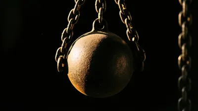 A heavy iron counterweight hangs still from a chain against black, one warm-lit rim of light tracing its rounded flank.