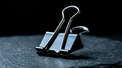 A single oversized black binder clip stands alone on dark slate, cold light tracing its upright folded handles.