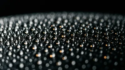 Hundreds of small water beads sit in an uneven scatter across a dark, gently curved matte surface, most catching a cold white pinpoint of light and some a warm amber one, the far rows blurring into black.