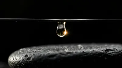 Una sola gota transparente cuelga del centro de un alambre fino que cruza el encuadre de borde a borde, con un punto de luz cálida brillando en su interior sobre la cúpula de una piedra oscura y picada sobre fondo negro.
