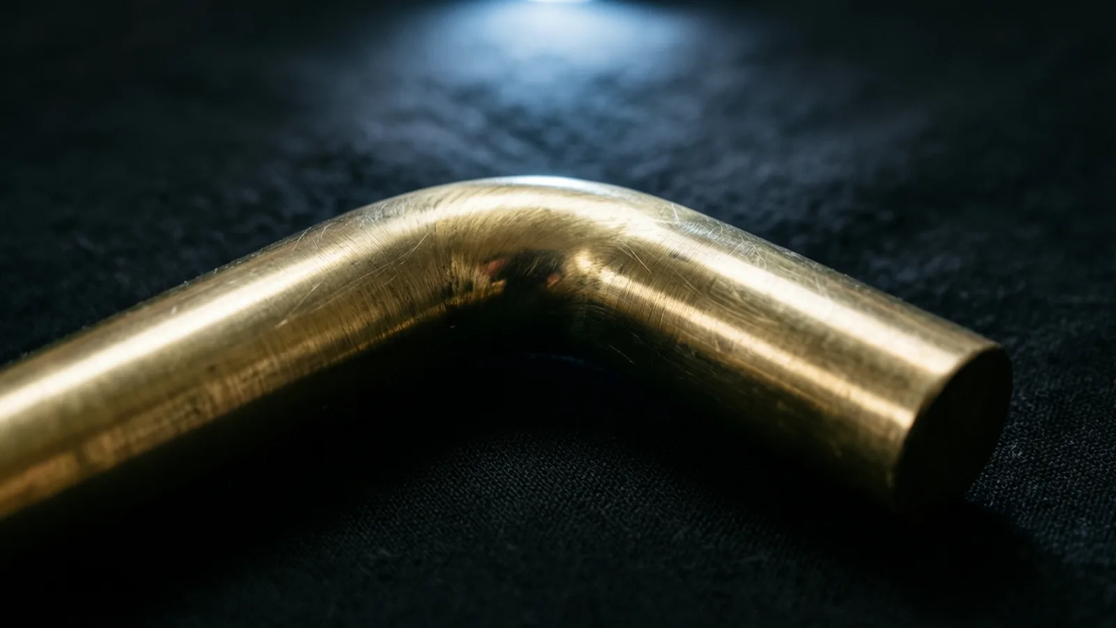 Una barra de metal doblada, con un brillo dorado y finos rayones, descansa sobre una tela oscura de trama visible, con su extremo cortado hacia la derecha. Un suave resplandor blanco azulado en la parte superior central ilumina la tela, y el fondo se funde en negro.