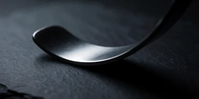 A single smooth curved sweep of brushed dark steel resting on dark slate, one bright arc of cold light tracing its curved edge against deep shadow.