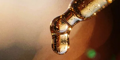 A single amber drop of viscous gel hanging from the tip of a glass pipette, lit from behind so the fine fibres suspended inside it catch the light against a warm out of focus ground.