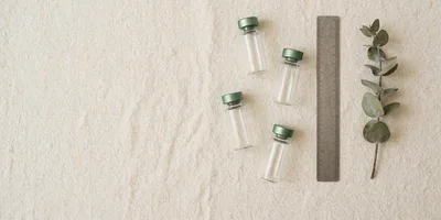 Four small glass vials with green anodised caps laid in a loose diagonal on cream linen, beside a brushed steel ruler and a single sprig of dried eucalyptus in soft overhead daylight.