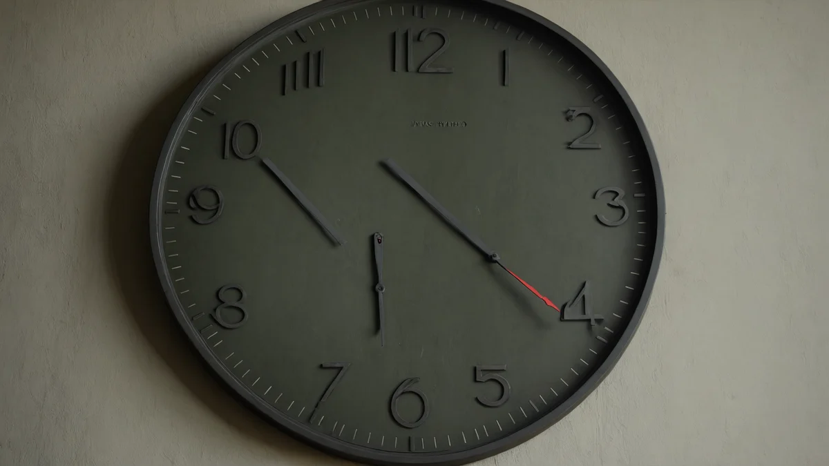 A plain grey-green wall clock seen at an angle in flat light, its slim hands the only detail on an otherwise empty wall.