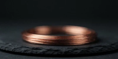 A neat coil of fine bare copper wire resting on dark slate, a single band of warm light catching the burnished loops so they glow against deep shadow.