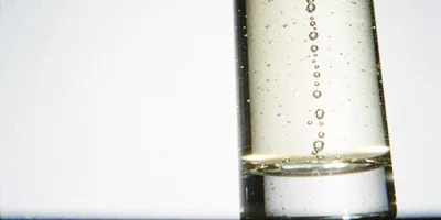 A tall glass column of pale straw coloured liquid backlit against a bright white ground so it glows, a single thread of fine bubbles rising through it.