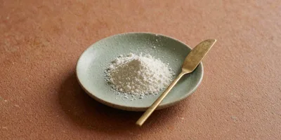 A small pile of fine white lyophilised powder in a pale sage ceramic dish on a warm terracotta surface, a slim brass spatula resting across the rim in soft afternoon light.