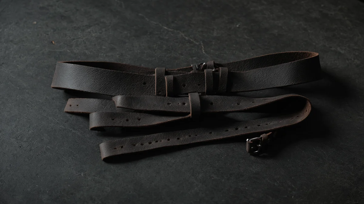 A worn dark leather strap coiled loosely on dark slate, its punched holes and finely cracked surface catching a low side light.