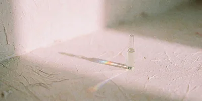 A single clear glass ampoule standing in soft blush pink light on a plaster surface, casting a long shadow with a faint prism of colour along its edge.