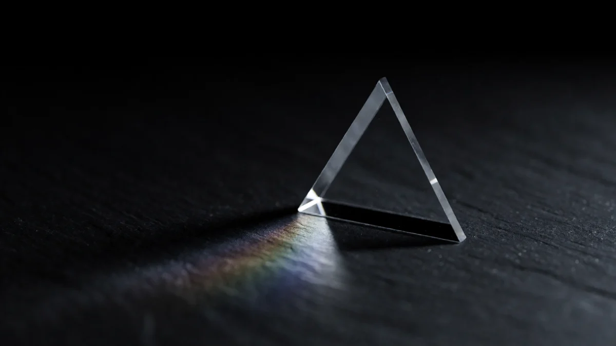 Un prisma triangular de vidrio transparente de pie sobre pizarra oscura, con una fina lamina de luz que lo atraviesa y se despliega en un tenue arcoiris sobre la piedra.