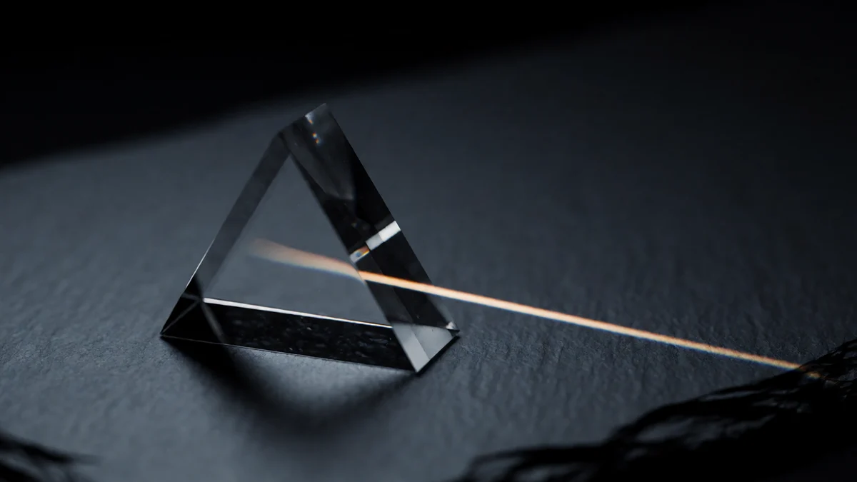 Un prisma triangular de vidrio macizo sobre pizarra oscura, con una fina lámina de luz cálida que lo atraviesa y se refracta en un tenue espectro.